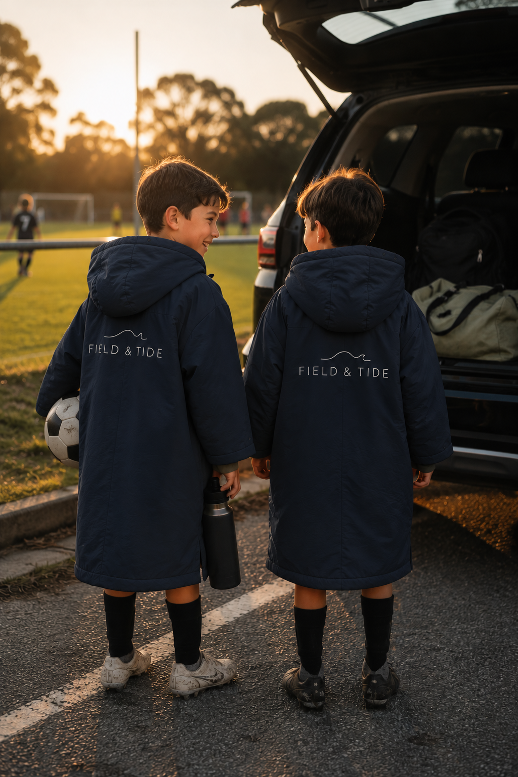 Two children in matching Field & Tide changing robes by an open car boot at a sports field at sunset